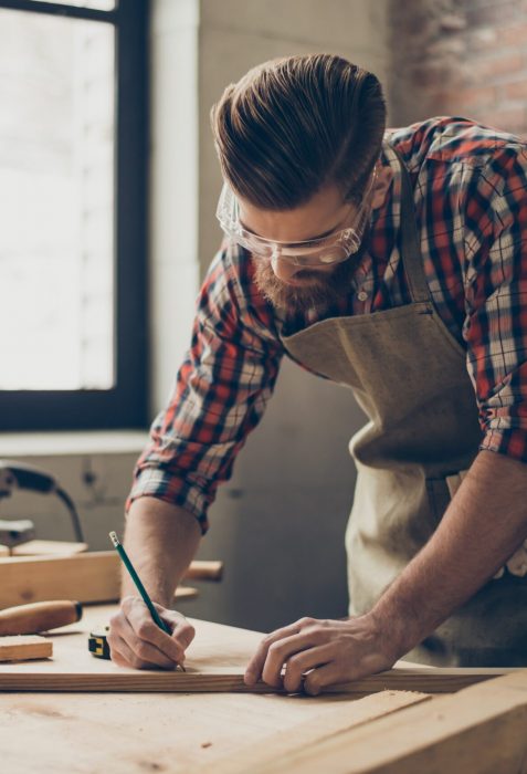 Bearded handsome cabinetmaker at the tabletop with pencil drawing sign on plank.  Stylish craftsman with brutal hairstyle and saved glasses work at his workstation.